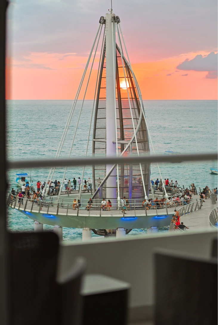 Pasarela al atardecer con gente, mar y cielo en tonos naranjas y rosados.