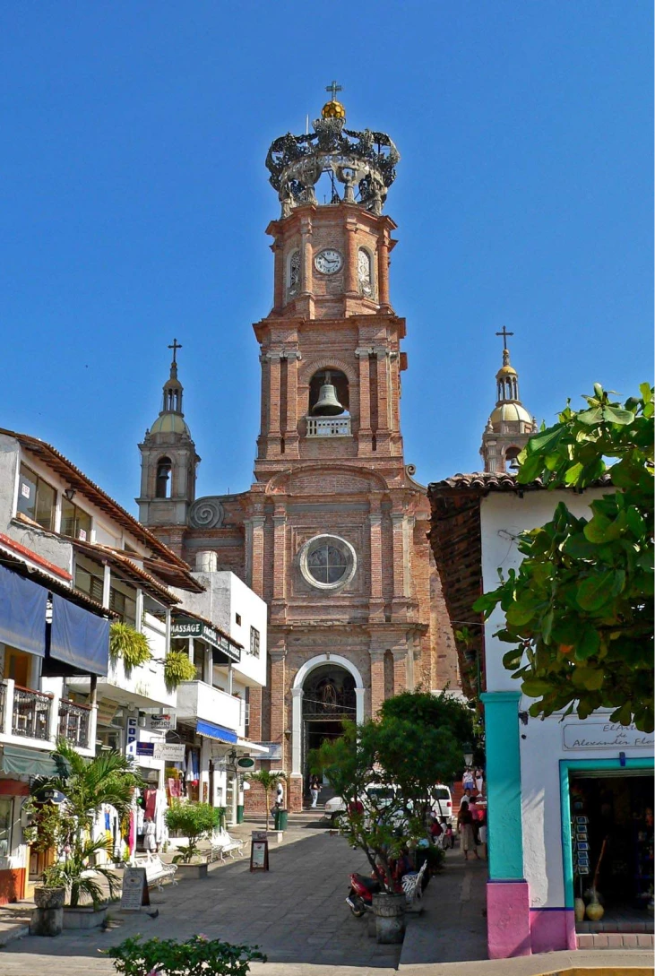 Iglesia de ladrillo con torre y corona en una calle soleada.