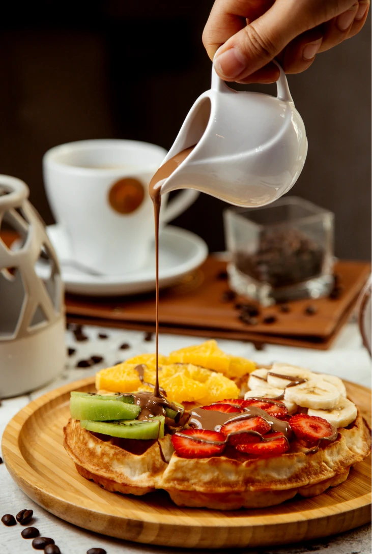 Waffle con frutas y chocolate l&iacute;quido, taza de caf&eacute; al fondo.