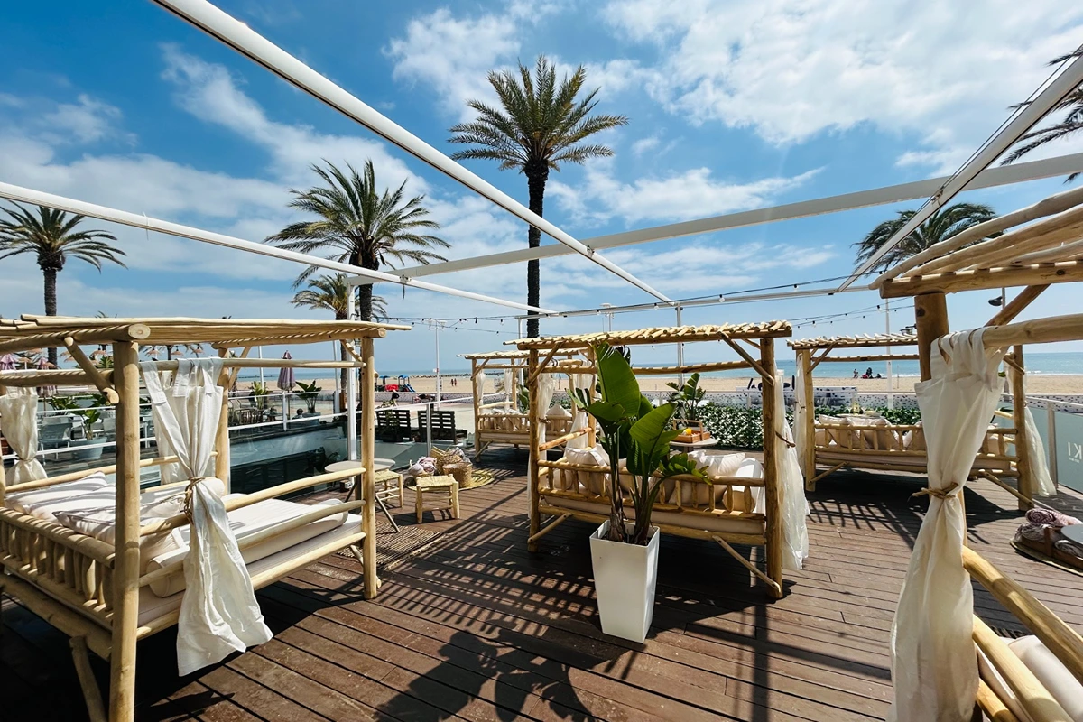 Terraza de playa con camas de bamb&uacute;, palmeras y cielo despejado.