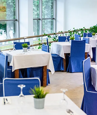 Restaurante con mesas cubiertas y sillas azules, plantas y ventanas grandes.