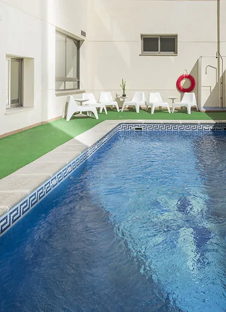 Piscina con sillas blancas, mesa y salvavidas colgado en la pared.