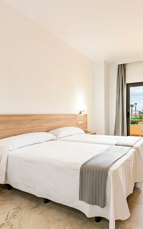 Habitaci&oacute;n luminosa con dos camas y un balc&oacute;n al fondo.