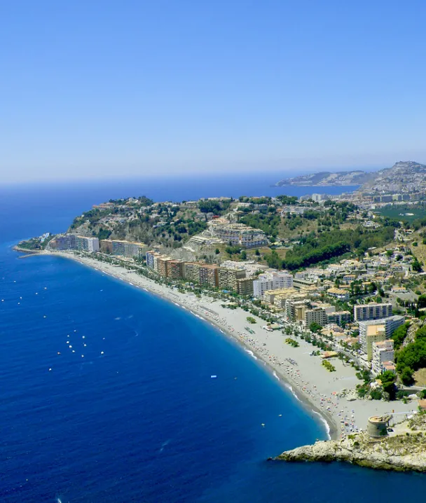 Costa panor&aacute;mica con playa, edificios y colinas verdes bajo un cielo despejado.
