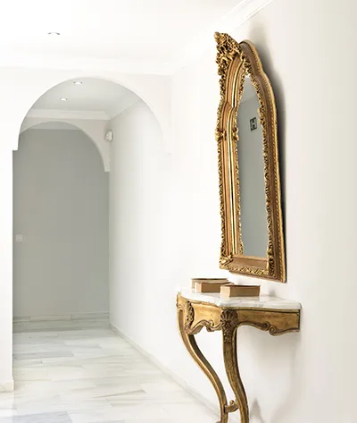 Pasillo blanco con espejo dorado decorativo sobre una mesa peque&ntilde;a.