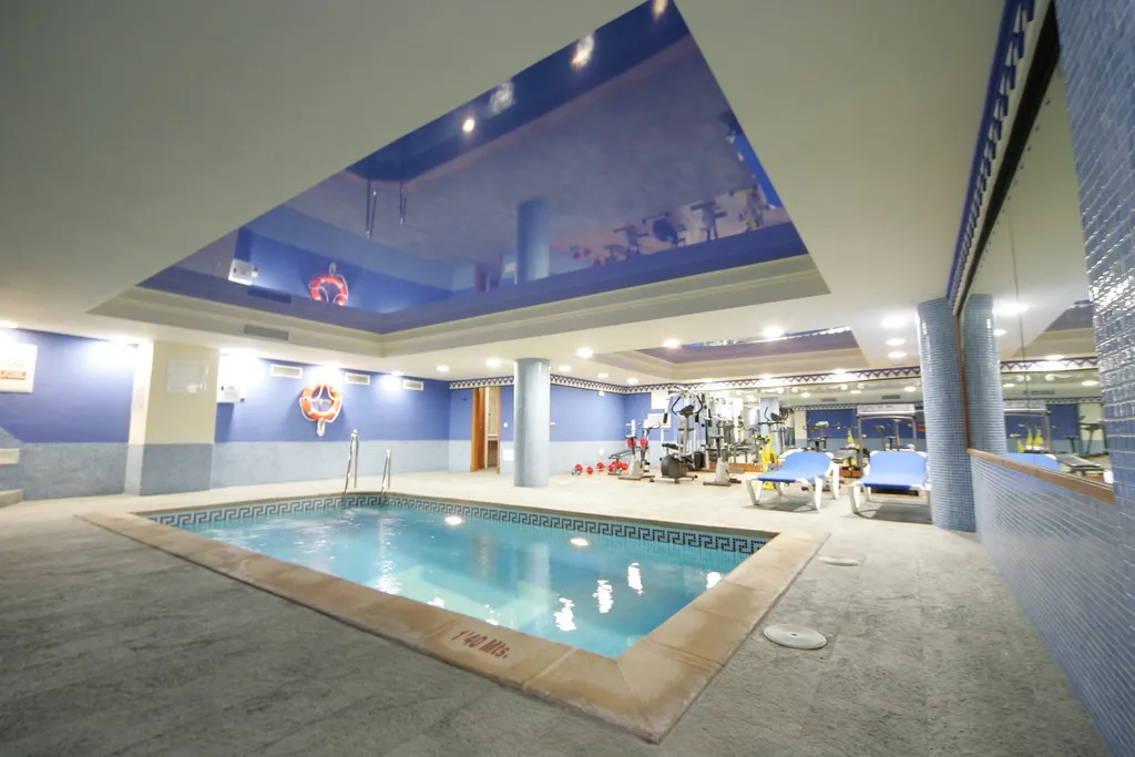 Piscina interior, gimnasio con equipo y tumbonas, decorado en azul y beige.