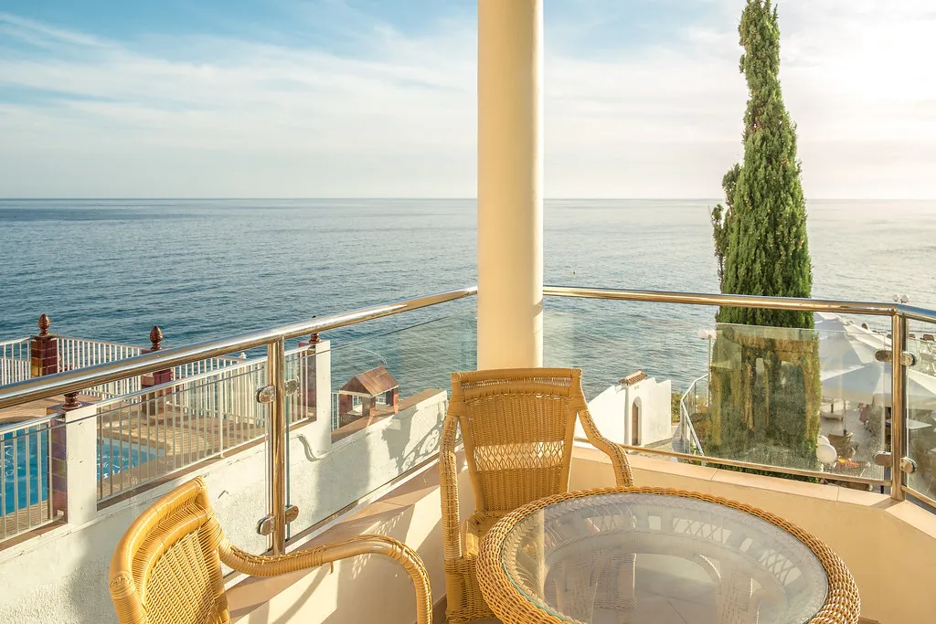 Balc&oacute;n con sillas y vista al mar, junto a una piscina y un &aacute;rbol alto.