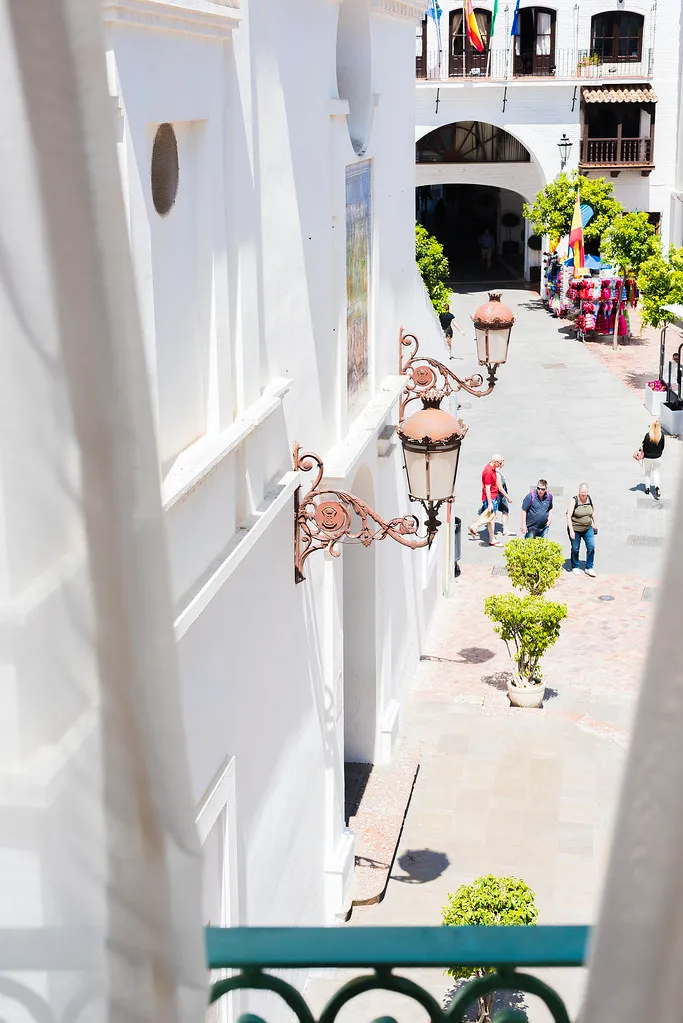 Vista desde una ventana a una calle con faroles y personas paseando.