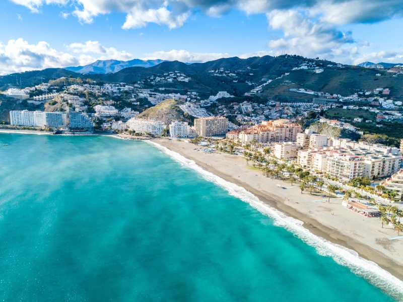 Vista a&eacute;rea de una playa con edificios y monta&ntilde;as al fondo bajo un cielo azul.