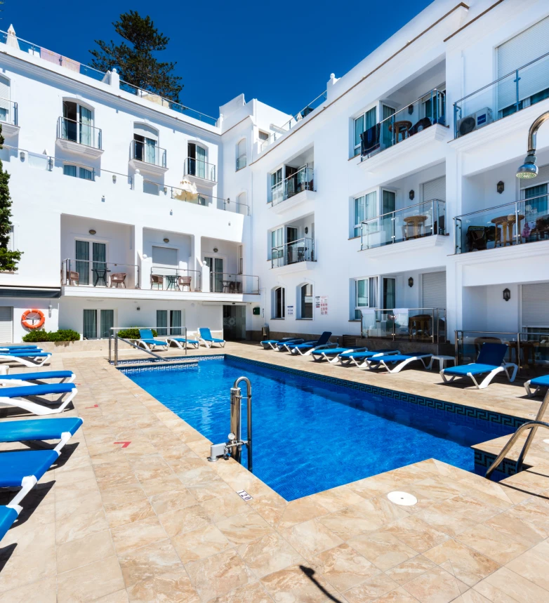 Piscina rodeada de tumbonas azules en un hotel blanco de varios pisos.