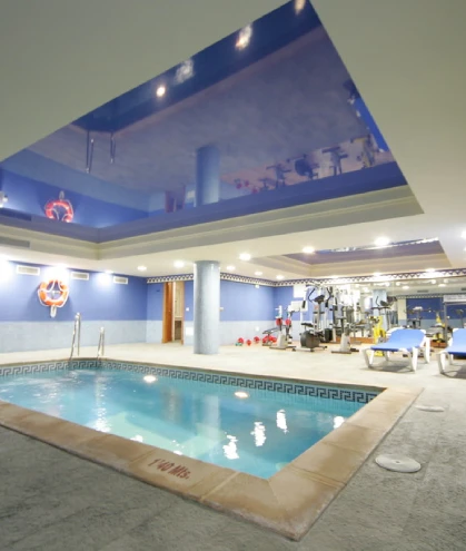 Piscina interior junto a un gimnasio con equipo y camillas azules.