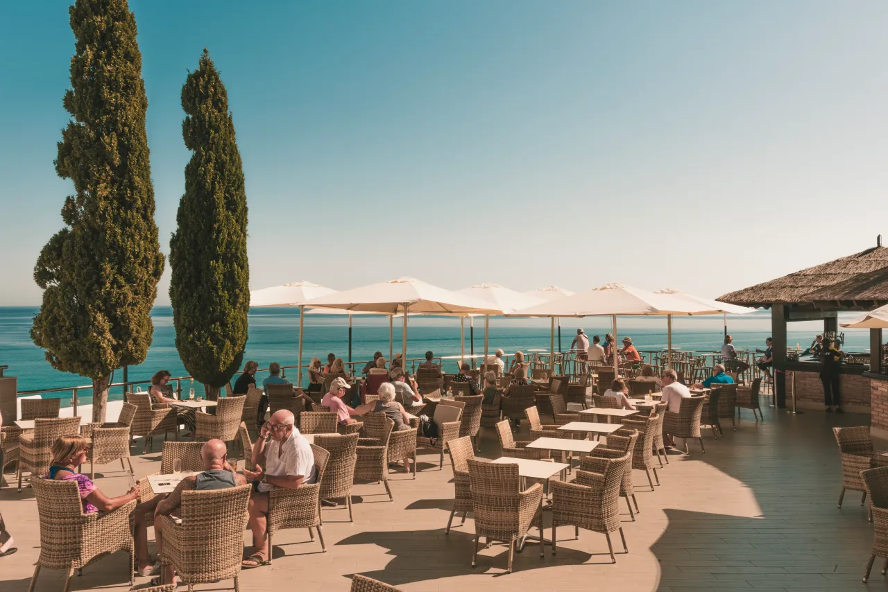 Personas disfrutando en una terraza con vistas al mar y sombrillas blancas.