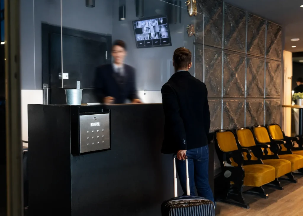 Hombre en recepci&oacute;n de hotel con maleta, interiores modernos, monitor arriba.