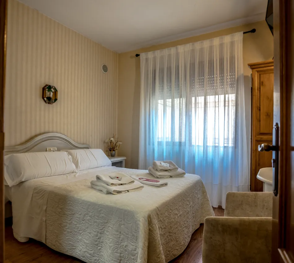 Dormitorio con cama doble, luz natural, muebles de madera y cortinas blancas.