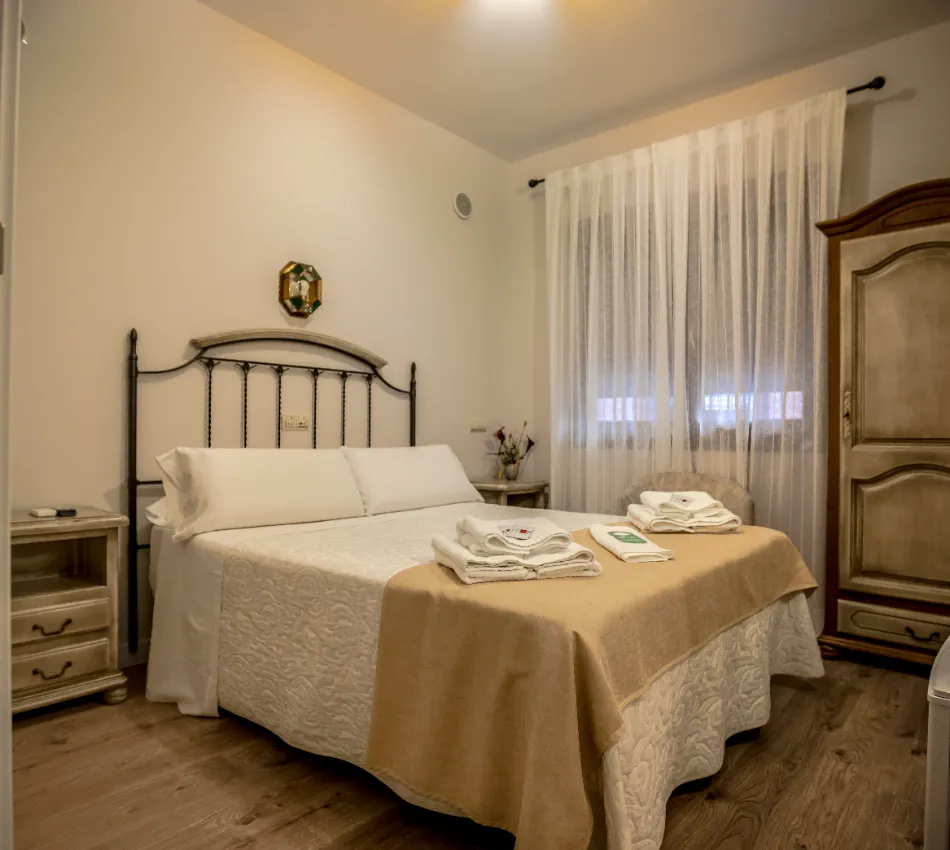 Habitaci&oacute;n acogedora con cama, mesitas y armario. Cortinas blancas y toallas sobre la cama.