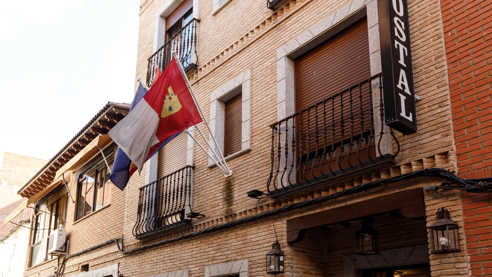 Fachada de hostal con banderas ondeando y ventanas con persianas marrones y barandales negros.