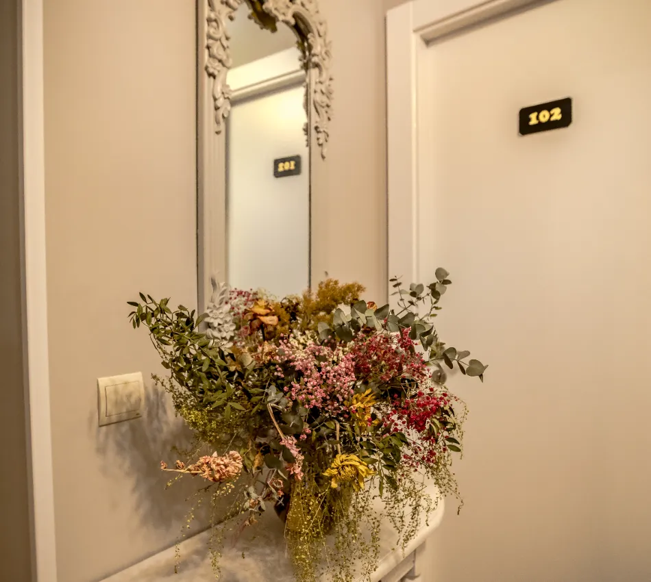 Ramo de flores frente a un espejo decorativo, junto a puerta blanca con n&uacute;mero 102.