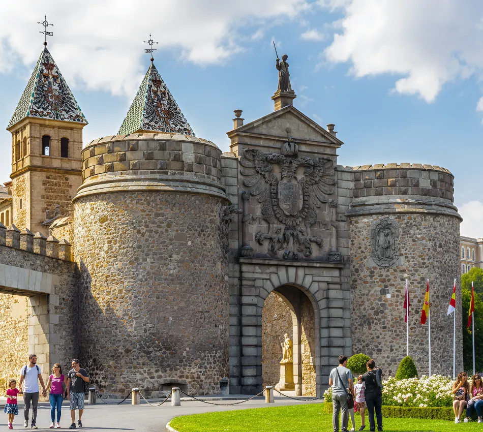 Antigua puerta de piedra con torres y escudos, pasando turistas en d&iacute;a soleado.