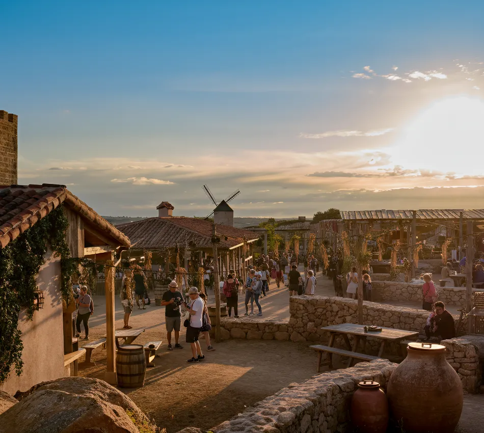Personas pasean por un pueblo con arquitectura r&uacute;stica al atardecer. Molinos al fondo.