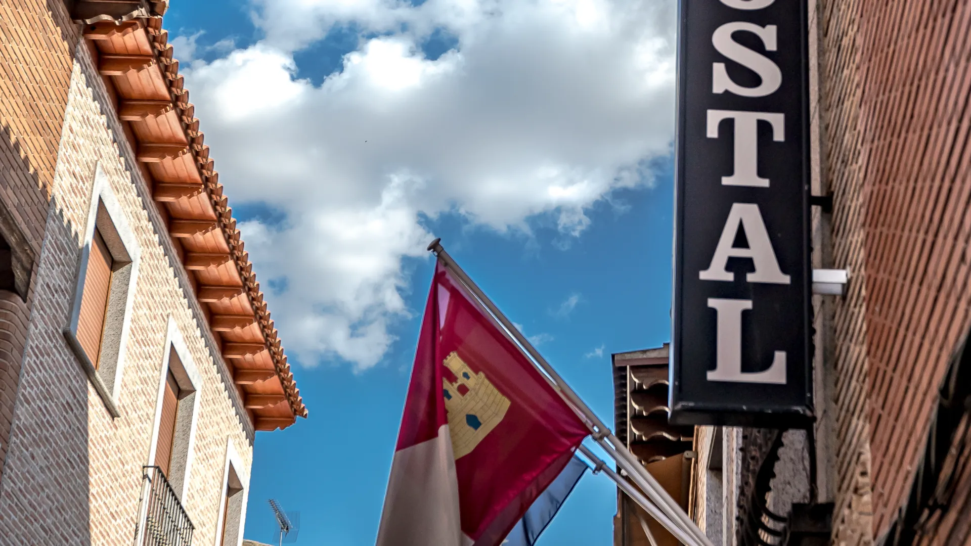 Edificios de ladrillo, bandera y cartel de "Hostal" bajo un cielo azul con nubes.