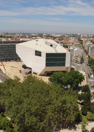 Edif&iacute;cio moderno em &aacute;rea urbana, rodeado por &aacute;rvores e ruas movimentadas.