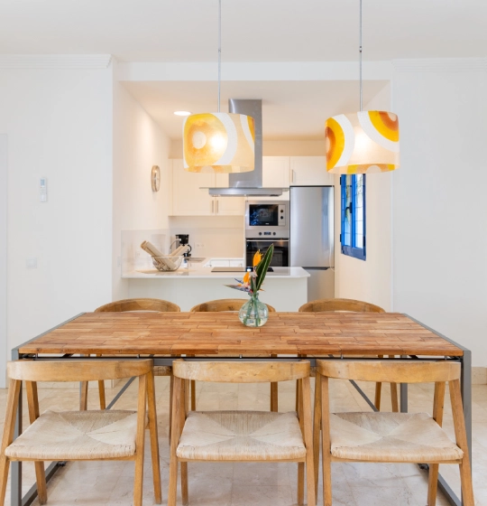 Comedor con mesa de madera, l&aacute;mparas colgantes y cocina de fondo.
