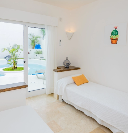 Habitaci&oacute;n blanca con cama, coj&iacute;n amarillo y vista a piscina y plantas.