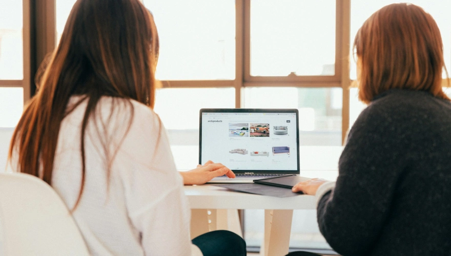 Dos personas usando una laptop en una mesa frente a una ventana.