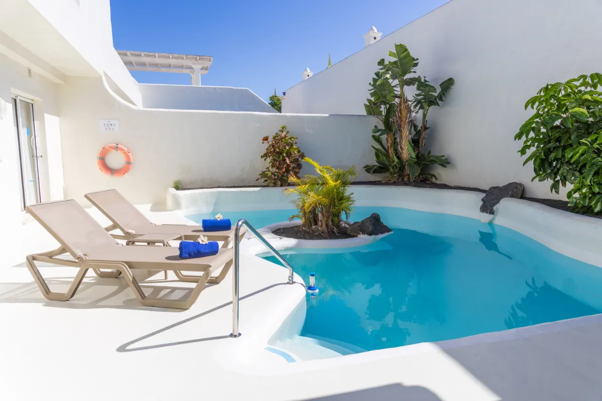 Piscina con tumbonas y plantas, rodeada por muros blancos bajo cielo azul.