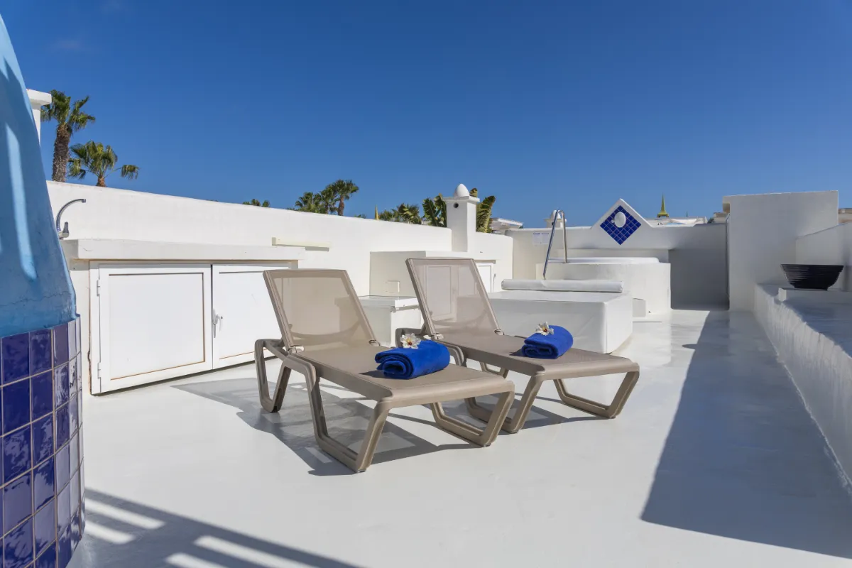 Tumbonas con toallas azules en terraza soleada y blanca bajo cielo azul.
