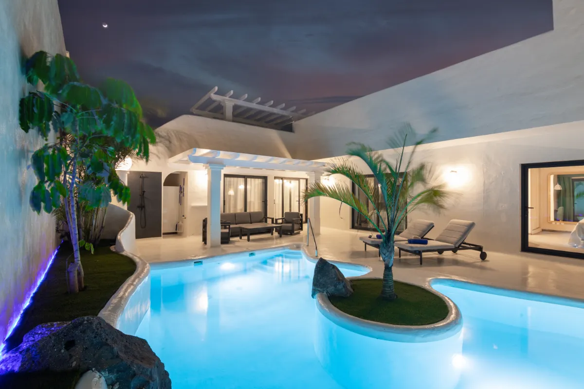 Piscina iluminada en una casa moderna con muebles de exterior y plantas.