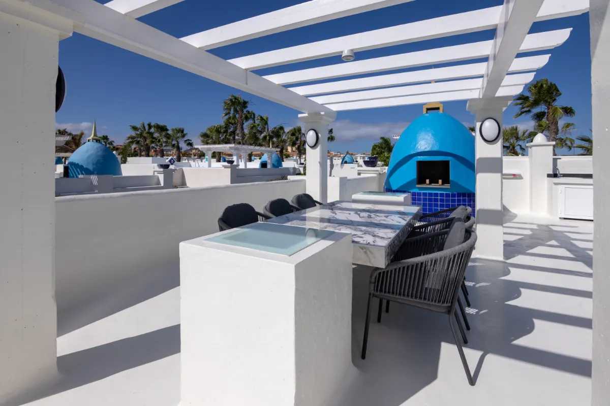 Terraza blanca con mesa, sillas y horno azul bajo un cielo despejado.