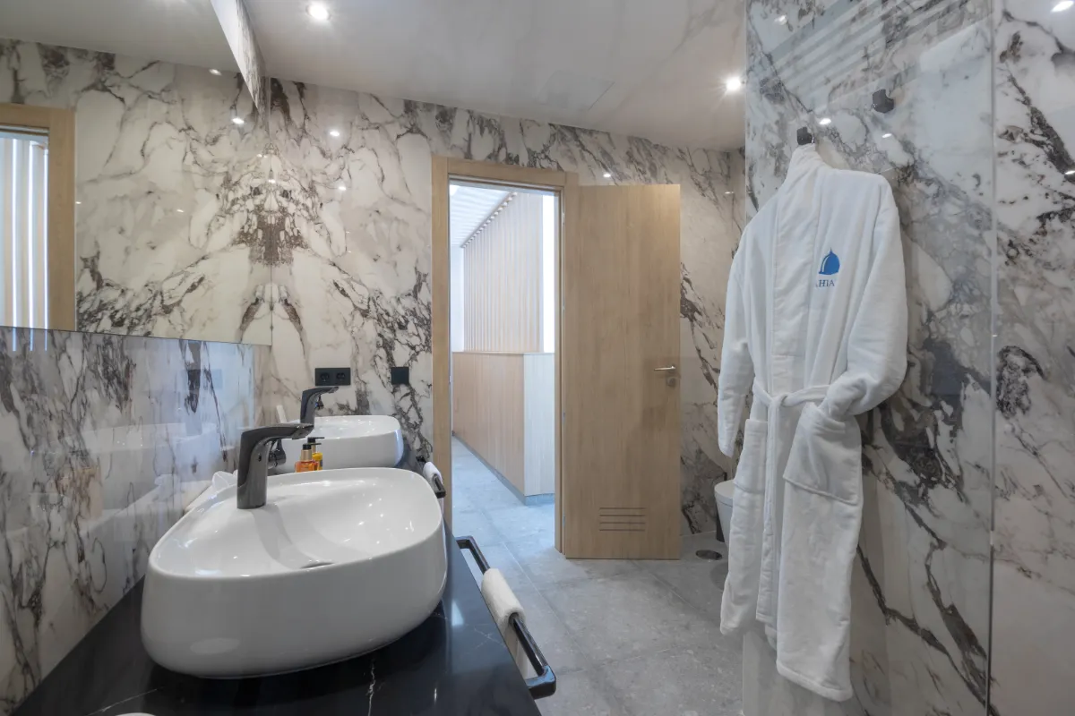 Baño moderno con lavabo doble y albornoz en pared de mármol.