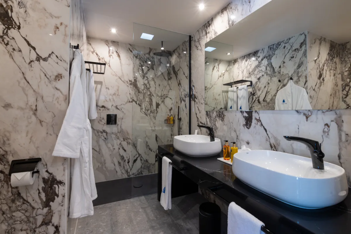 Baño moderno con lavabos blancos, paredes de mármol y espejo grande.
