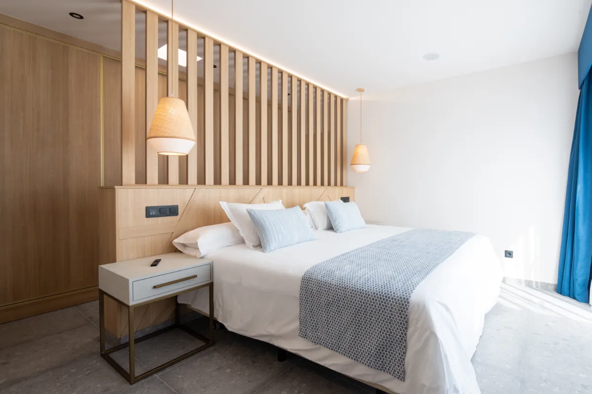 Cama con cojines grises, lámpara colgante y pared de madera decorativa.