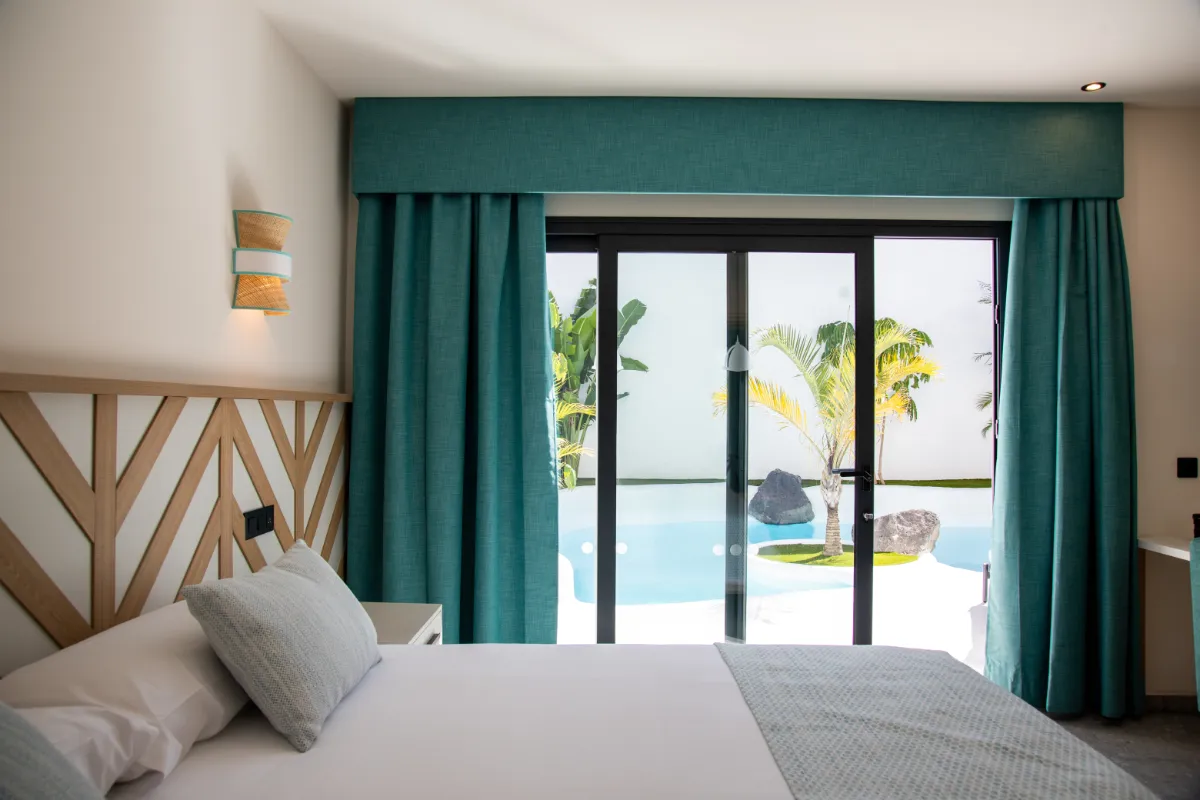 Habitación luminosa con cama, cortinas turquesa y vista a la piscina y palmeras.