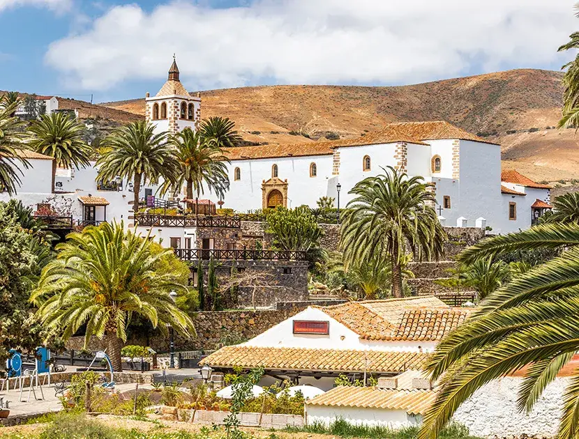 Pueblo pintoresco con iglesia, palmeras y colinas al fondo.