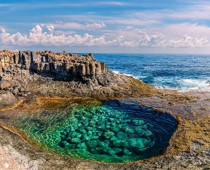 Charco de agua cristalina sobre rocas volc&aacute;nicas junto al oc&eacute;ano bajo un cielo azul.