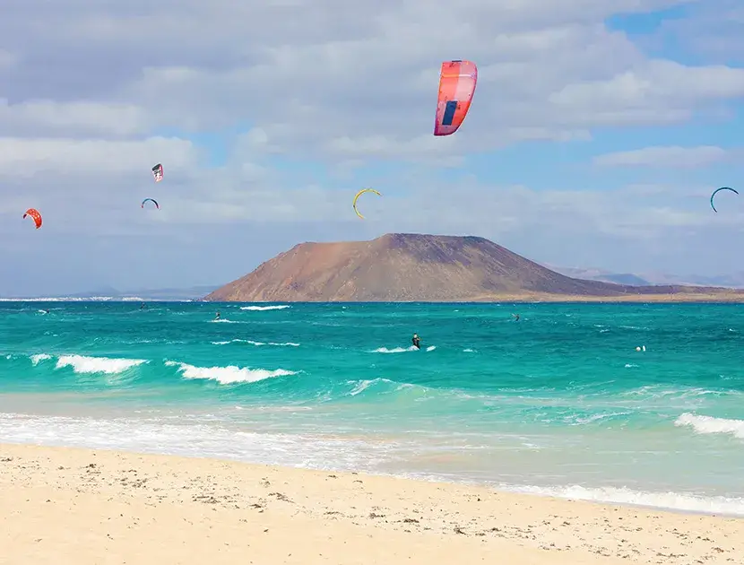 Playa con cometas de kitesurf y una isla monta&ntilde;osa al fondo.