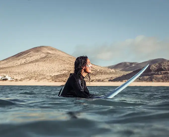Persona surfea en el mar con colinas des&eacute;rticas al fondo.