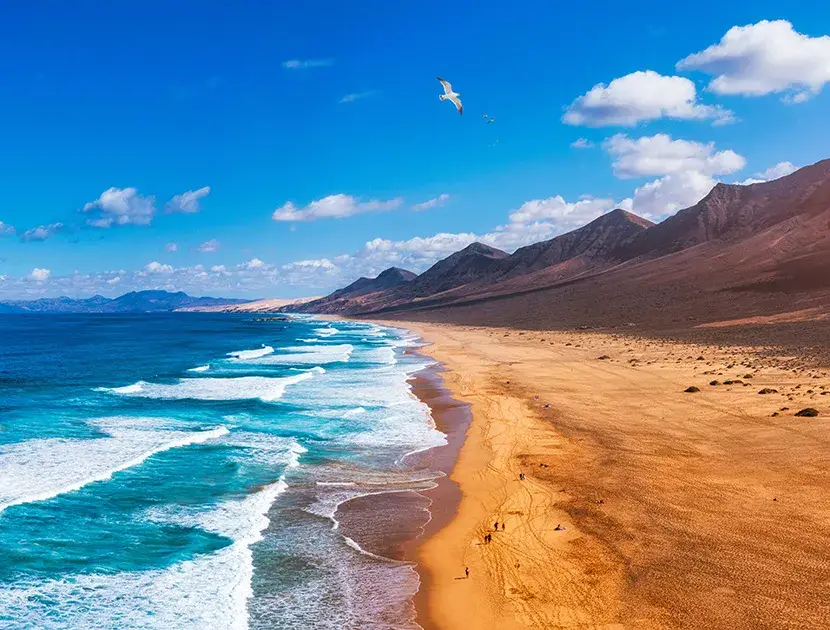 Playa amplia con olas, monta&ntilde;as y cielo azul con nubes dispersas.