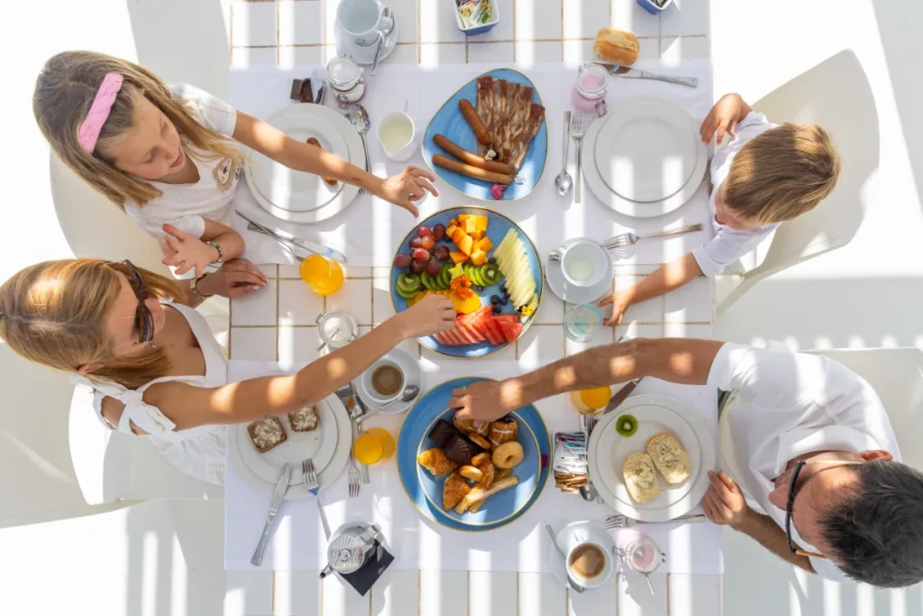 Familia disfrutando de un desayuno variado al aire libre en una mesa blanca.