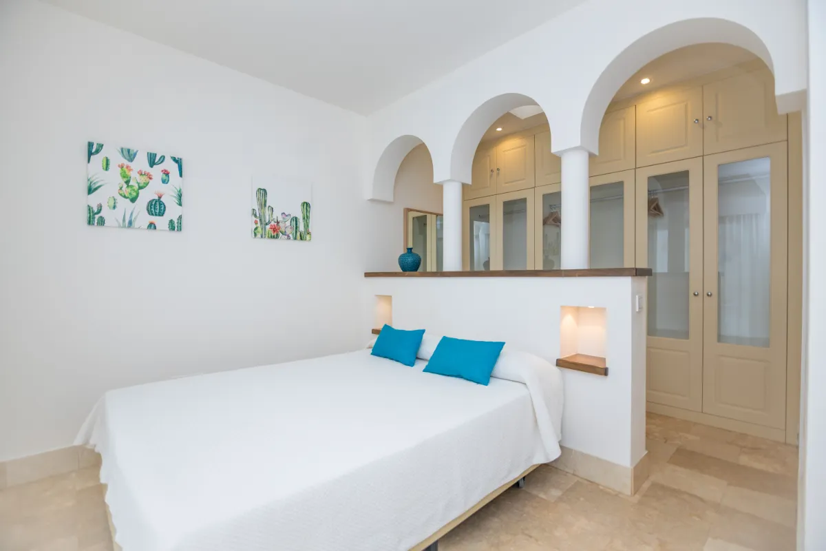 Habitación con cama blanca, cojines azules y decoración de cactus en la pared.
