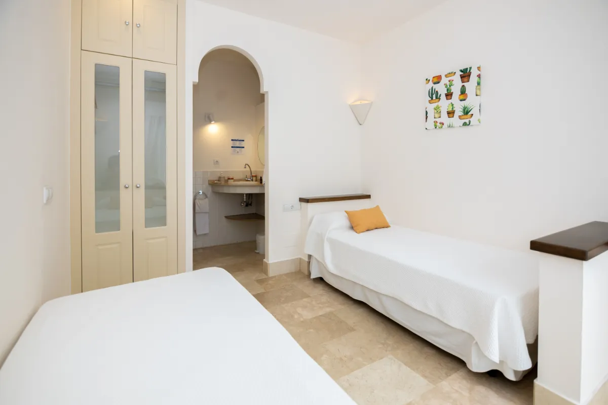Habitación blanca con dos camas y un baño al fondo.