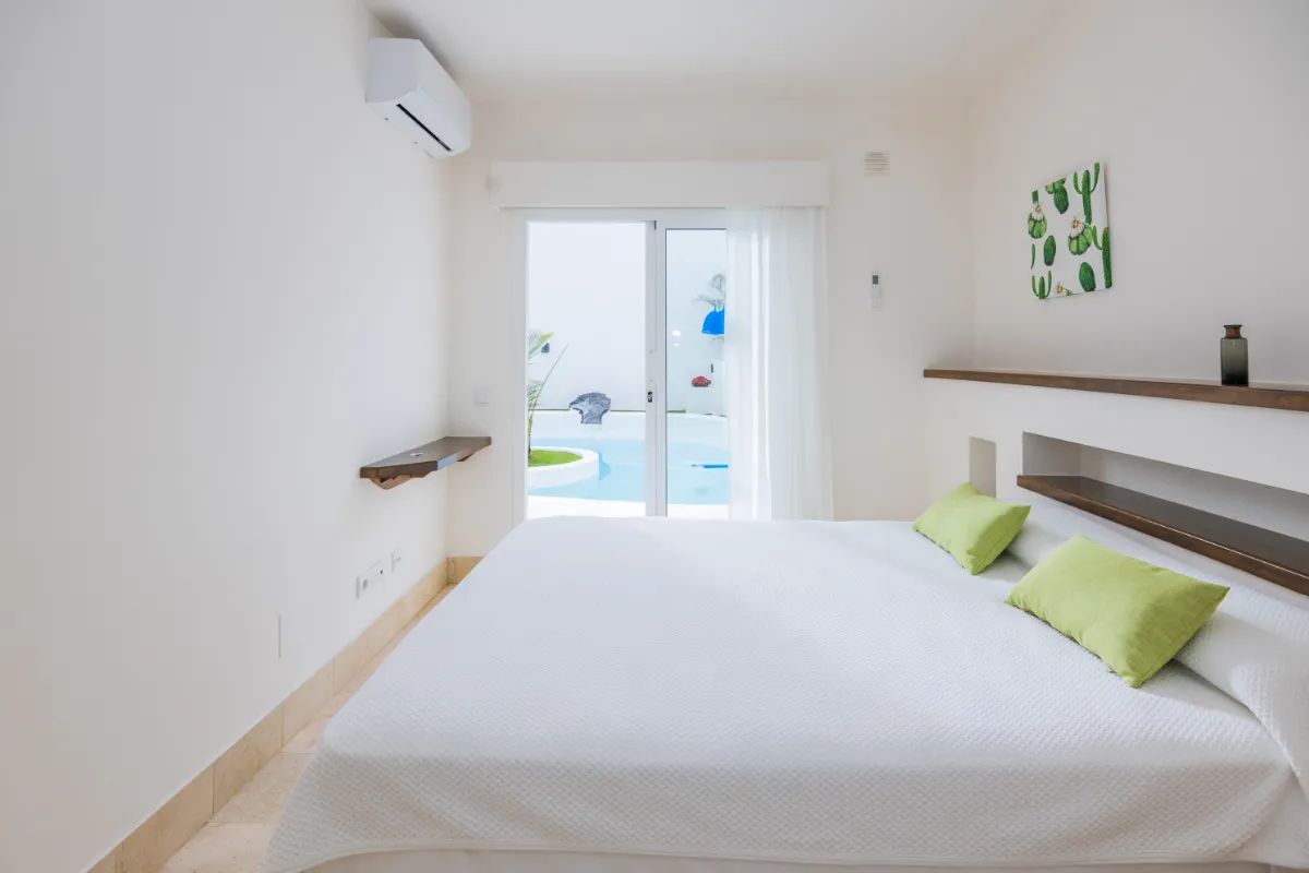 Habitación blanca con cama, cojines verdes y vista a la piscina exterior.