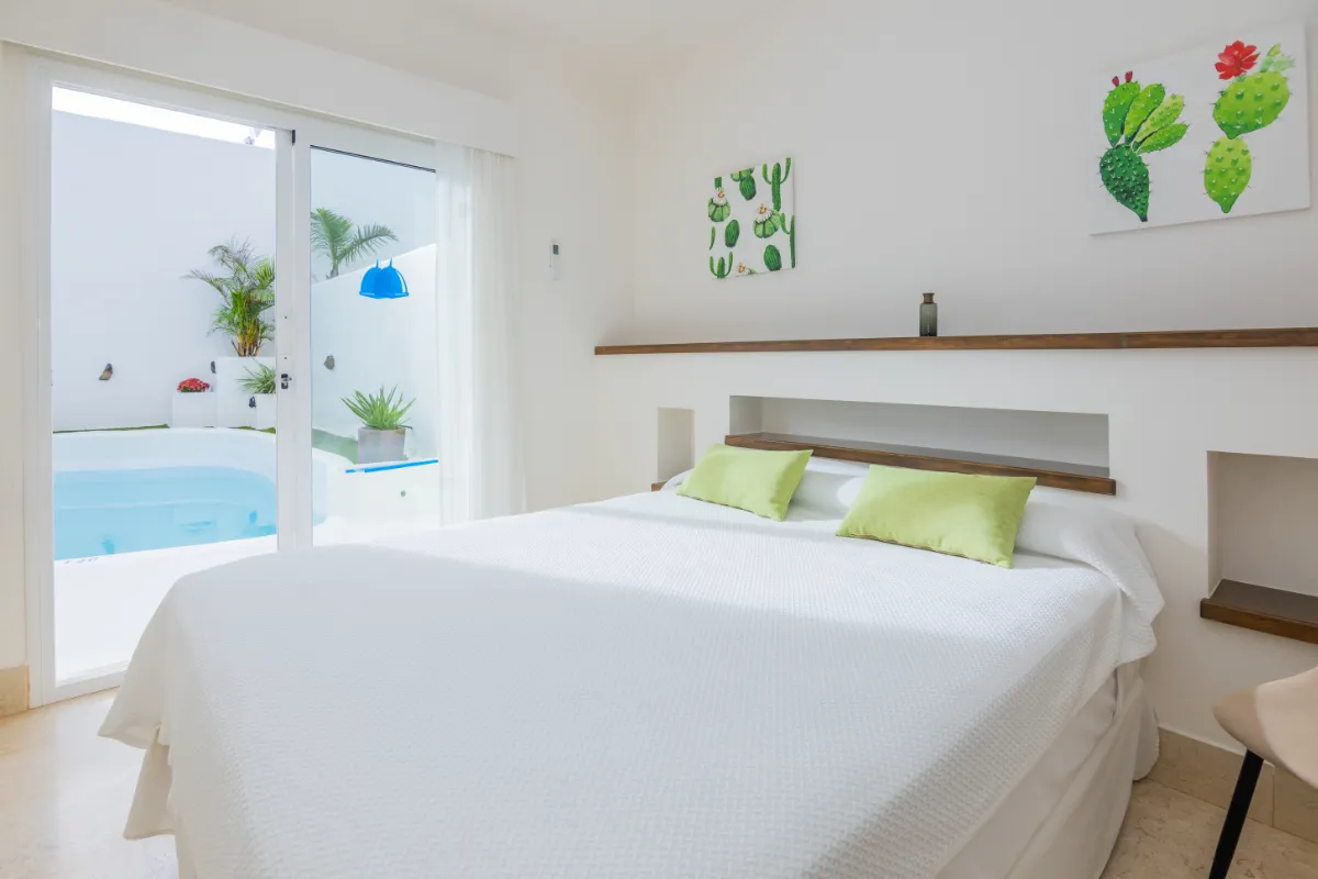 Habitación con cama blanca, cojines verdes y vista a una piscina exterior.