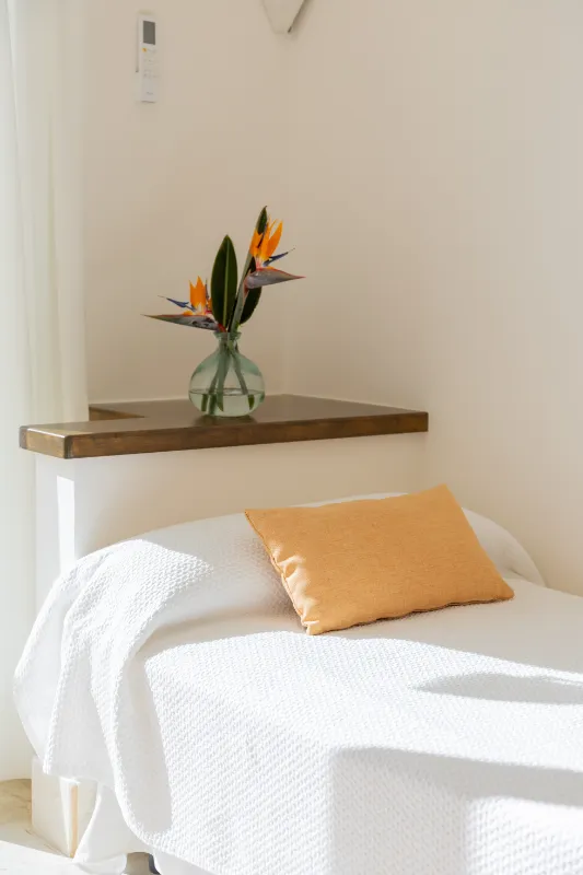 Cama blanca con almohada amarilla y flores en jarr&oacute;n sobre mueble.