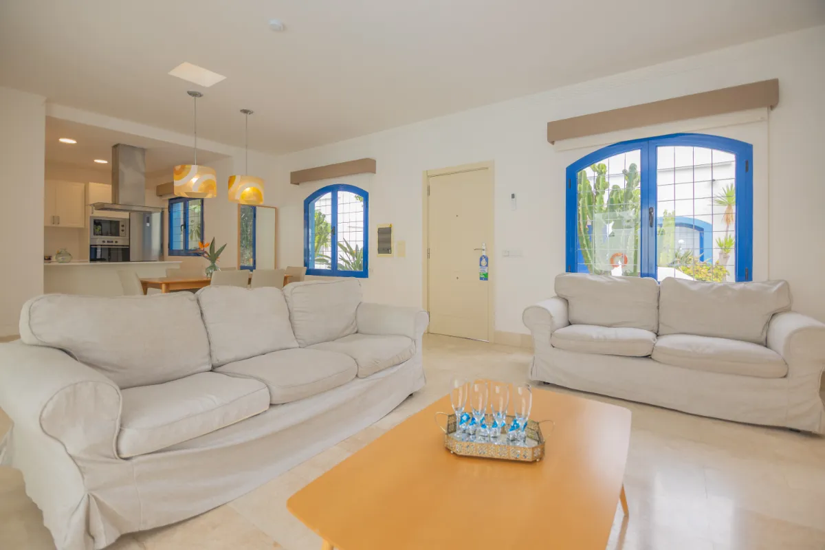 Sala de estar con sofás blancos, mesa de centro y ventanas con marcos azules.
