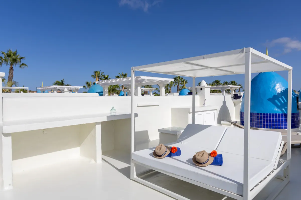 Cama blanca con sombreros en una terraza soleada y cielo azul.