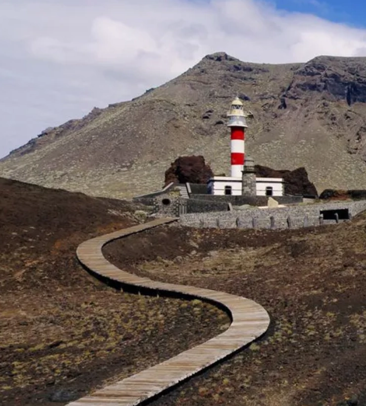 Camino sinuoso hacia un faro con fondo de monta&ntilde;a rocosa y cielo parcialmente nublado.